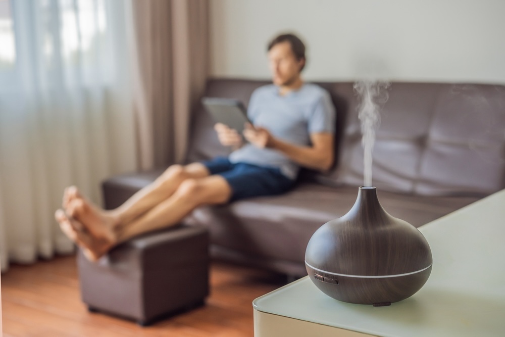 A man enjoys the calming vapor from an essential oil diffuser while relaxing with a tablet in his modern living room.